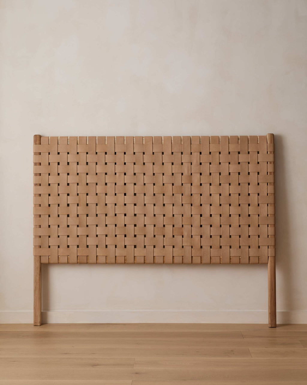 A light woven leather headboard with a woven basketweave pattern and matching wooden legs, set against a neutral beige wall and light wood floor.