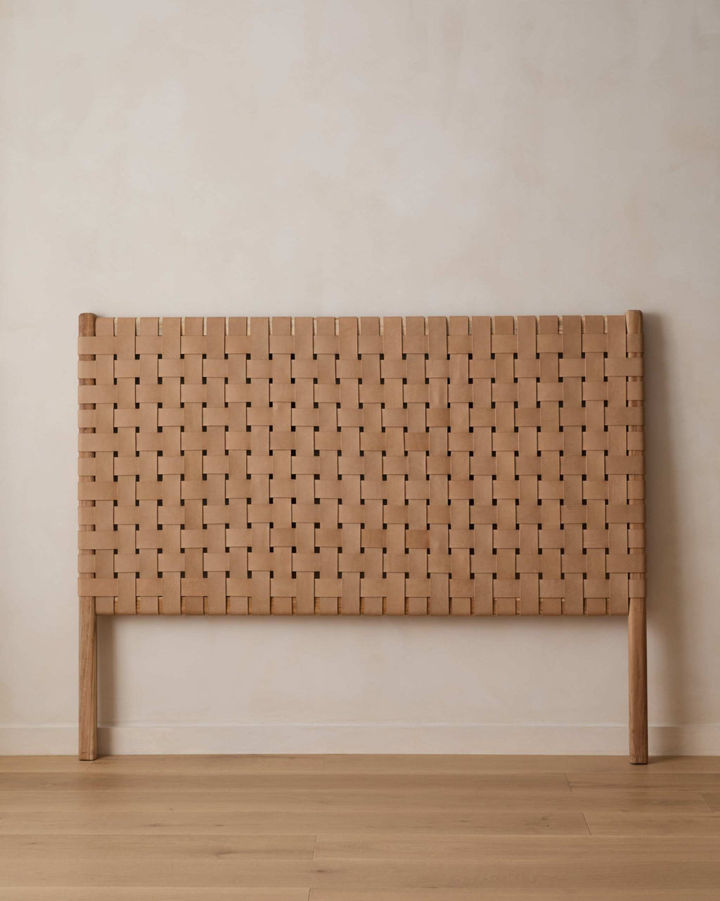 A light natural woven leather headboard with a woven basketweave pattern and matching wooden legs, set against a neutral beige wall and light wood floor.