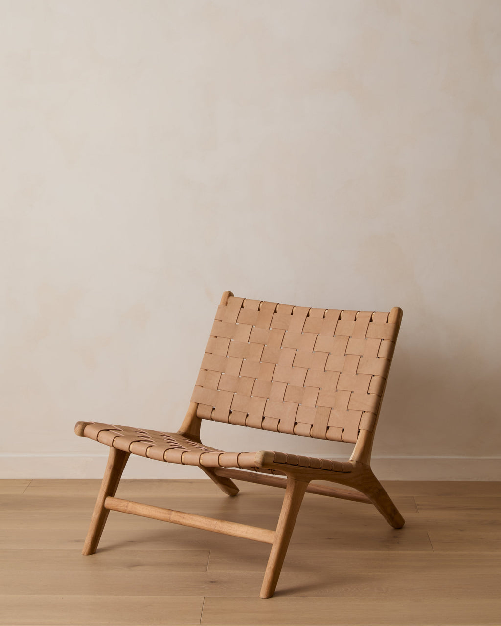 A woven leather lounge chair with a woven tan leather seat and backrest, set against a minimalist beige wall and light wood floor.