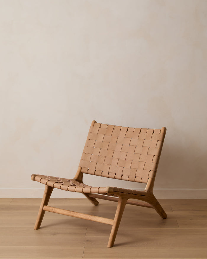A woven leather lounge chair with a woven tan leather seat and backrest, set against a minimalist beige wall and light wood floor.