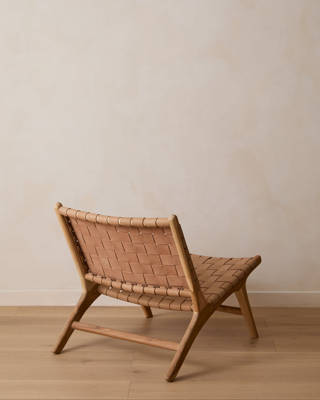 A woven leather lounge chair with woven tan leather straps, shown from a low angle against a neutral beige wall and light wood floor.