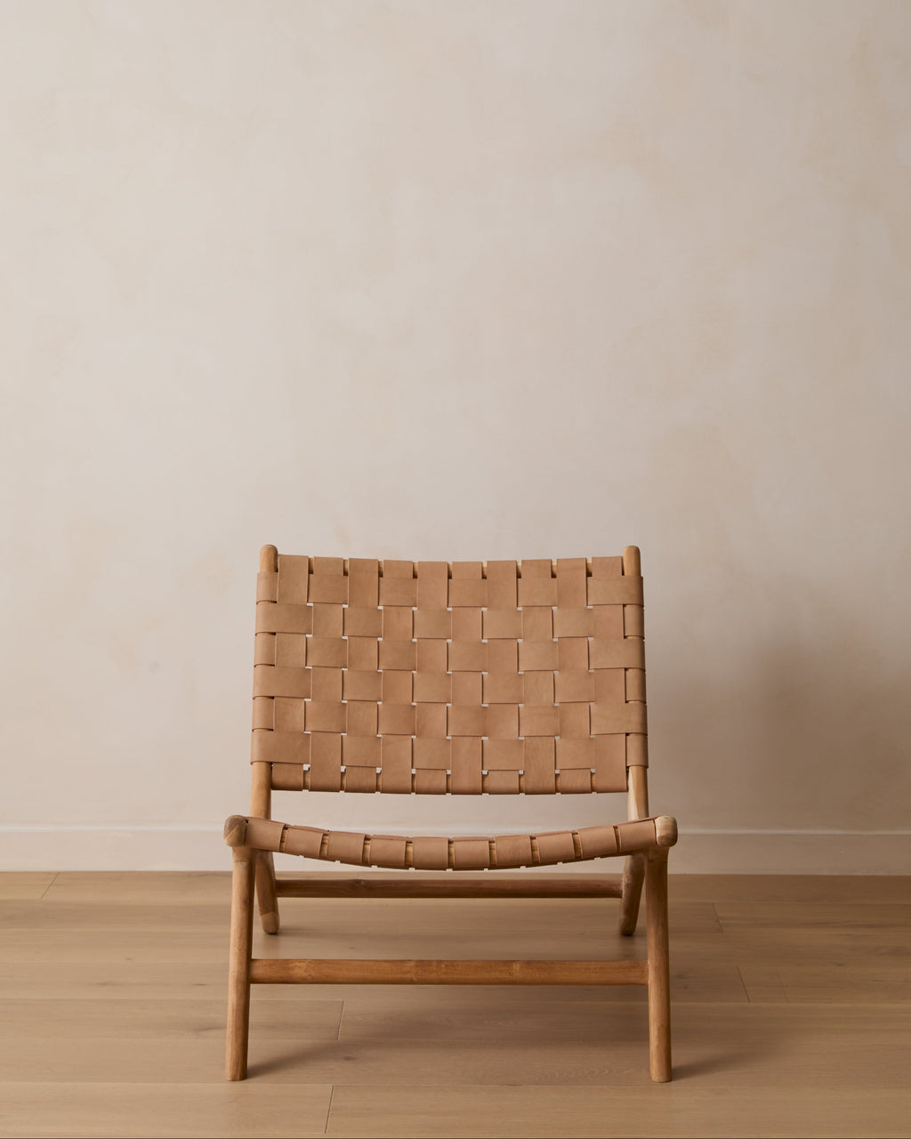A woven leather lounge chair with a woven tan leather seat and backrest on a light wood frame, set against a minimalist beige wall and light wood floor.