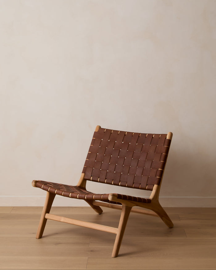 A woven leather lounge chair with a saddle woven leather seat and backrest, set against a minimalist beige wall and light wood floor.