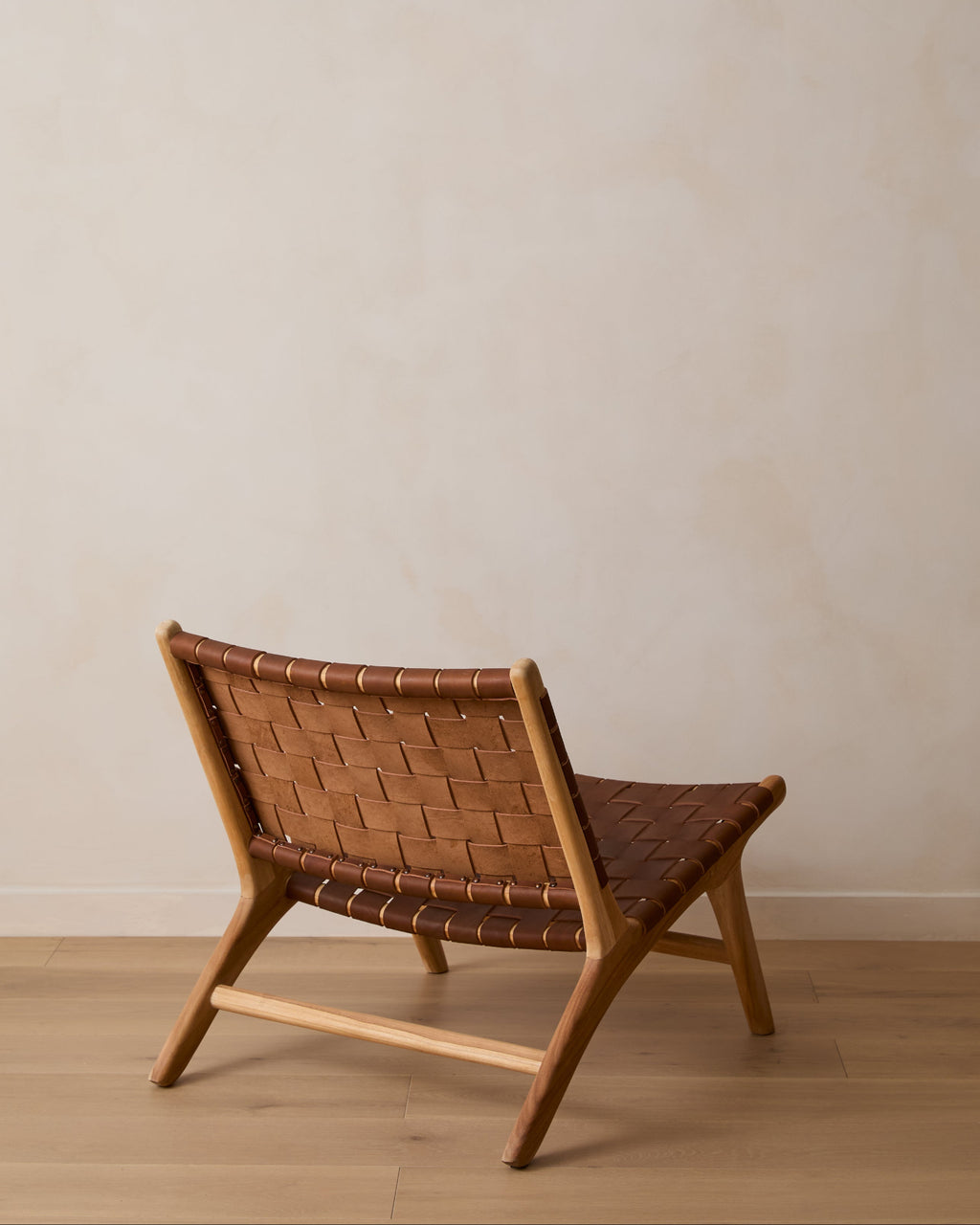 A woven leather lounge chair with a woven saddle leather seat and backrest, positioned on a light wood floor against a textured beige wall.