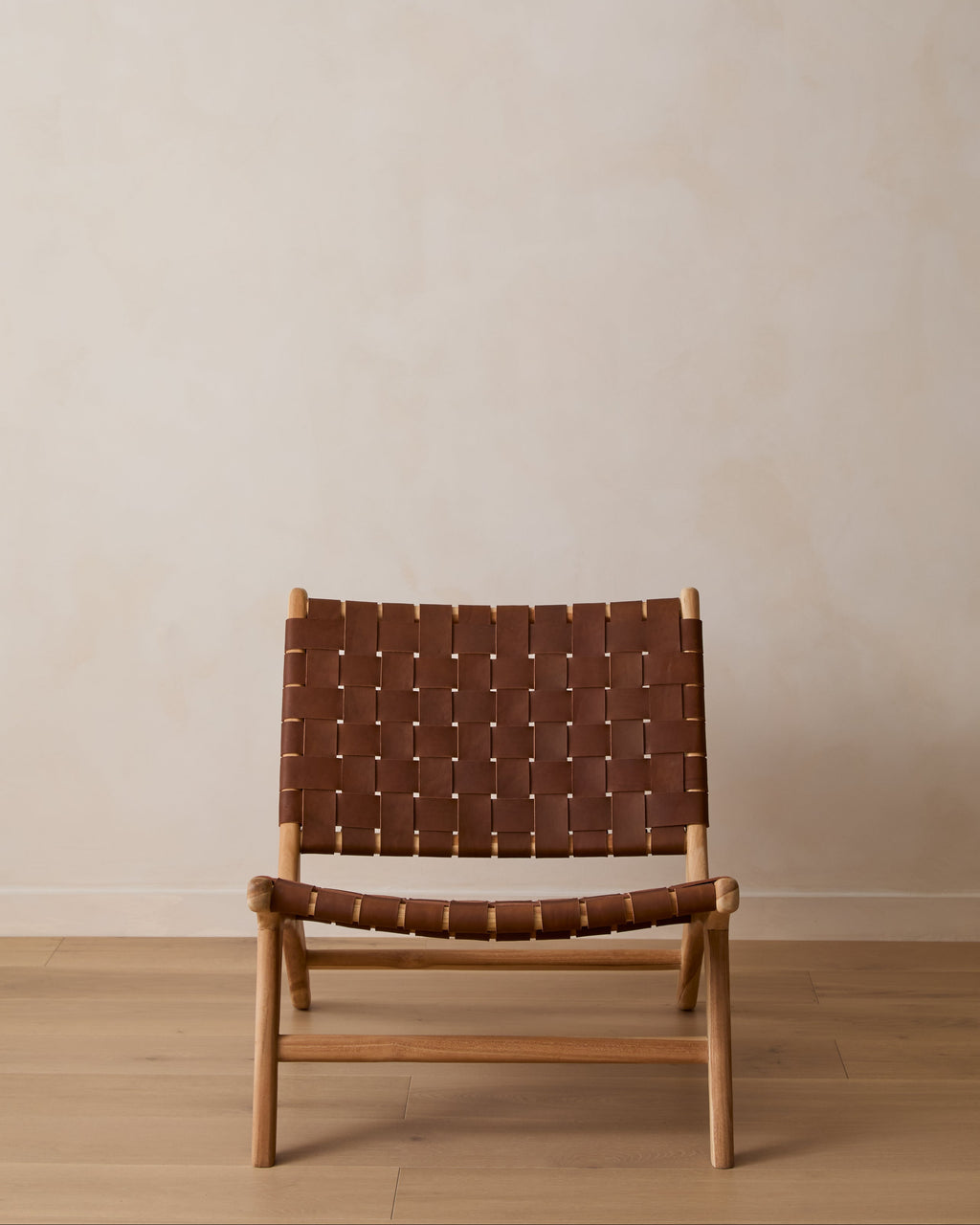 A woven leather lounge chair with a saddle leather woven sling seat and back, set against a minimalist beige wall and light wood floor.