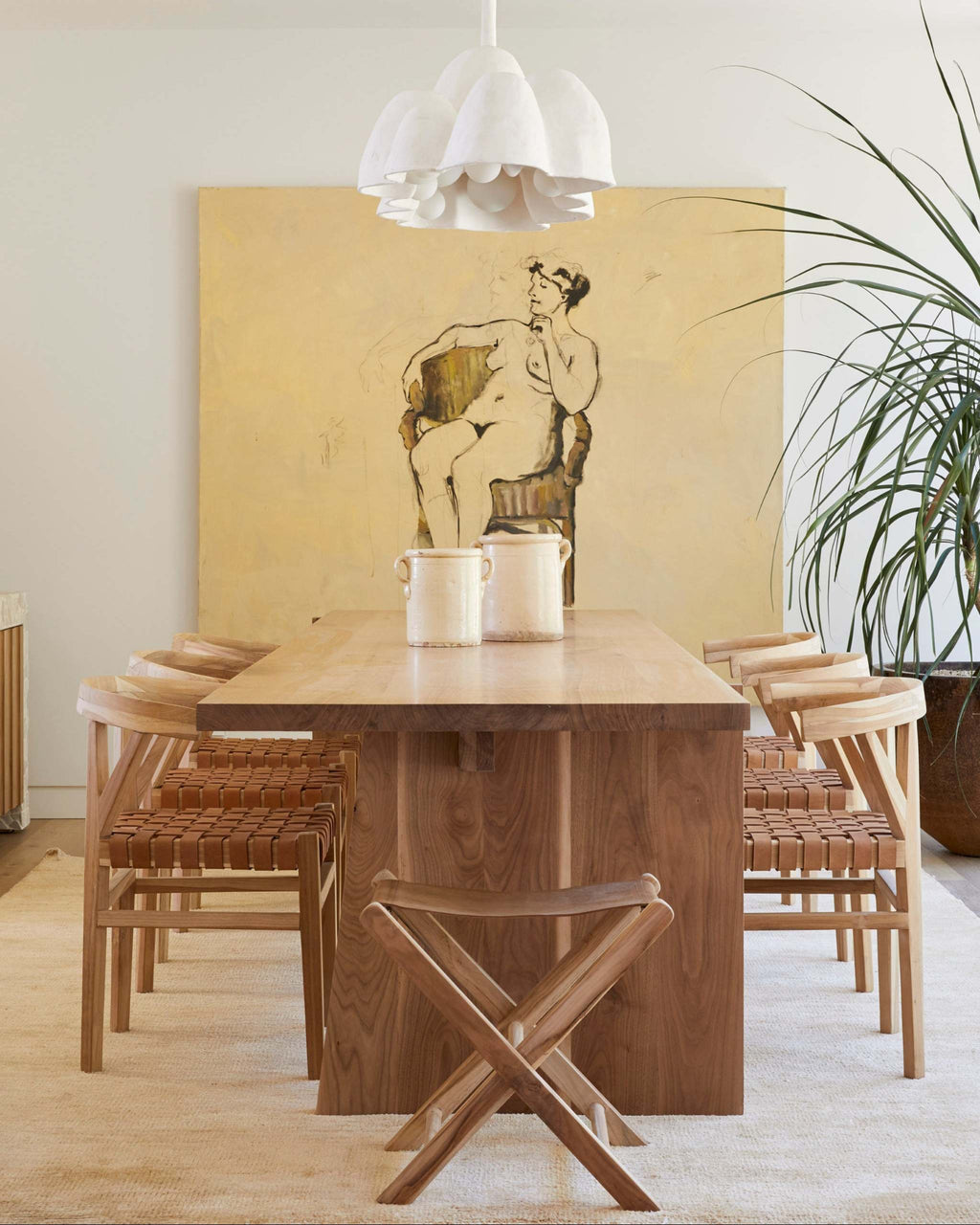 A modern dining room setup featuring a rectangular wooden dining table surrounded by six wishbone woven leather dining chairs, under a white multi-tiered pendant light. A large yellow canvas painting with a black line drawing of a seated figure hangs on the wall behind the table.