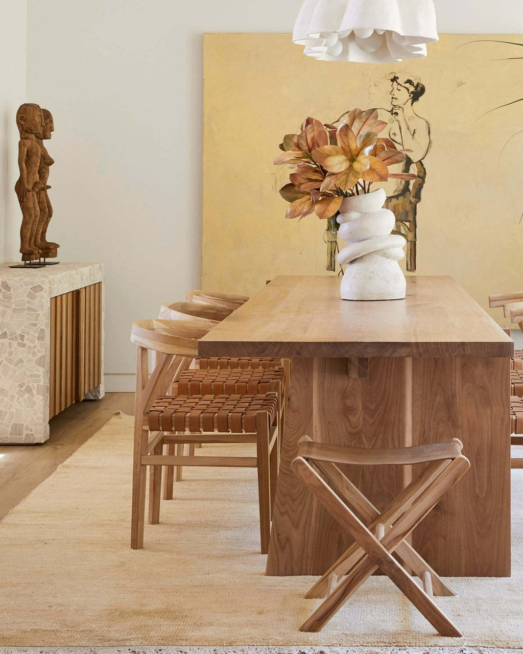 A warm, modern dining room scene featuring a rectangular wooden dining table, woven leather wishbone dining chairs, a small wooden folding stool, and a white sculptural vase with dried flowers on the table, set against a large yellow abstract painting.
