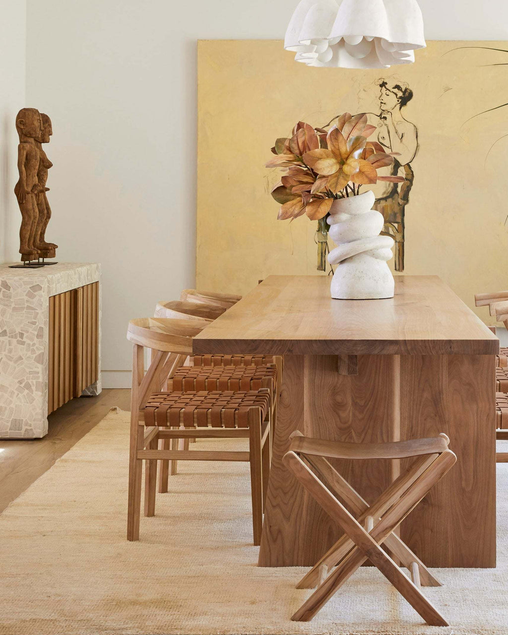 A warm, modern dining room scene featuring a rectangular wooden dining table, woven leather wishbone dining chairs, a small wooden folding stool, and a white sculptural vase with dried flowers on the table, set against a large yellow abstract painting.