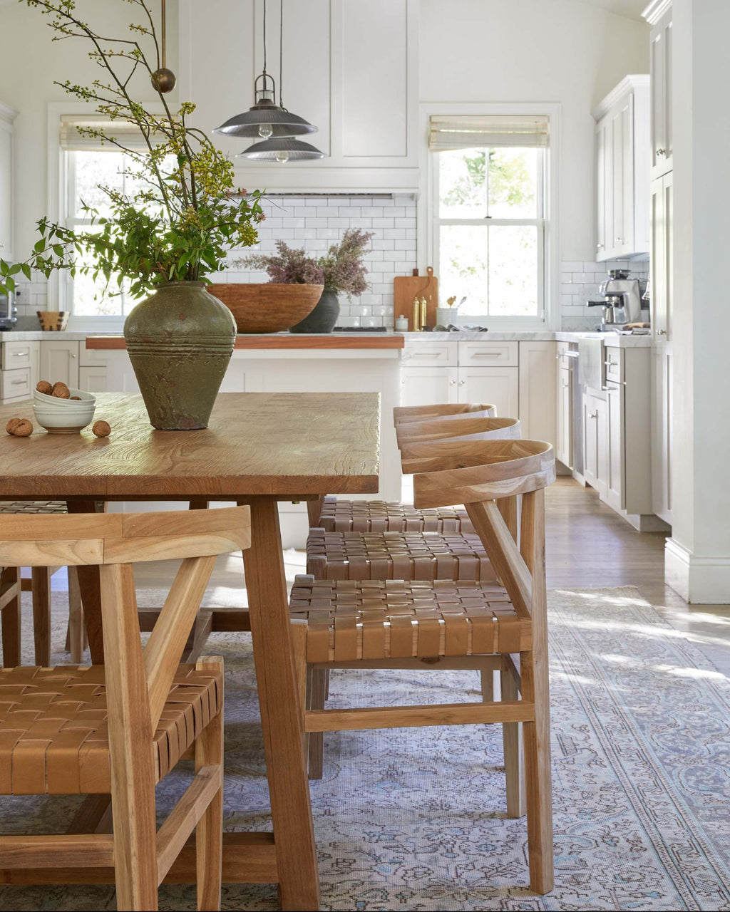 A bright, modern kitchen with white cabinetry, a large wooden dining table, woven leather wishbone dining chairs, and a tall green ceramic vase filled with greenery in the foreground.