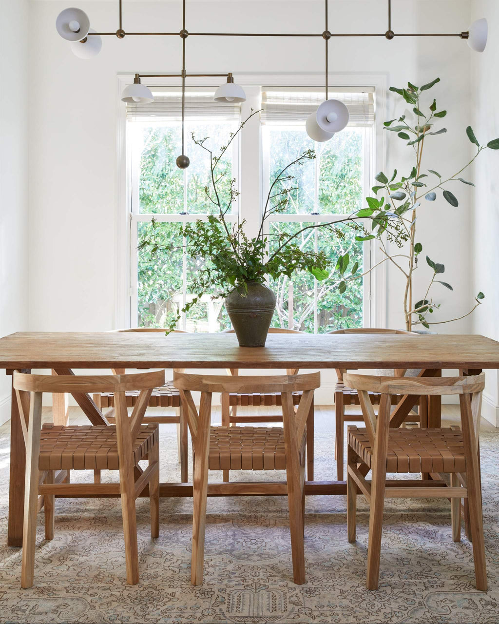 A modern dining room scene featuring a light oak wood dining table and six woven leather wishbone dining chairs with woven seats, set on a patterned rug in front of a large window.