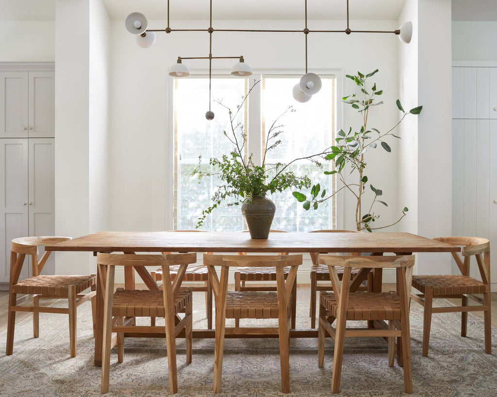 A modern dining room setup featuring a rectangular light oak wood table and six matching woven leather wishbone dining chairs with woven cane seats, arranged on a textured gray rug in front of a large window.