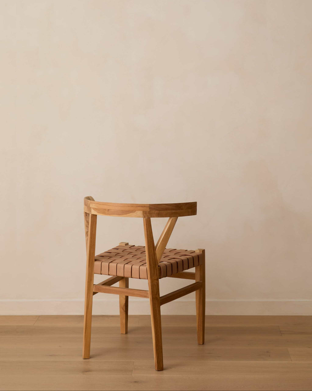 A woven leather wishbone dining chair with a woven seat strap, positioned against a neutral beige plaster wall and light wood floor.