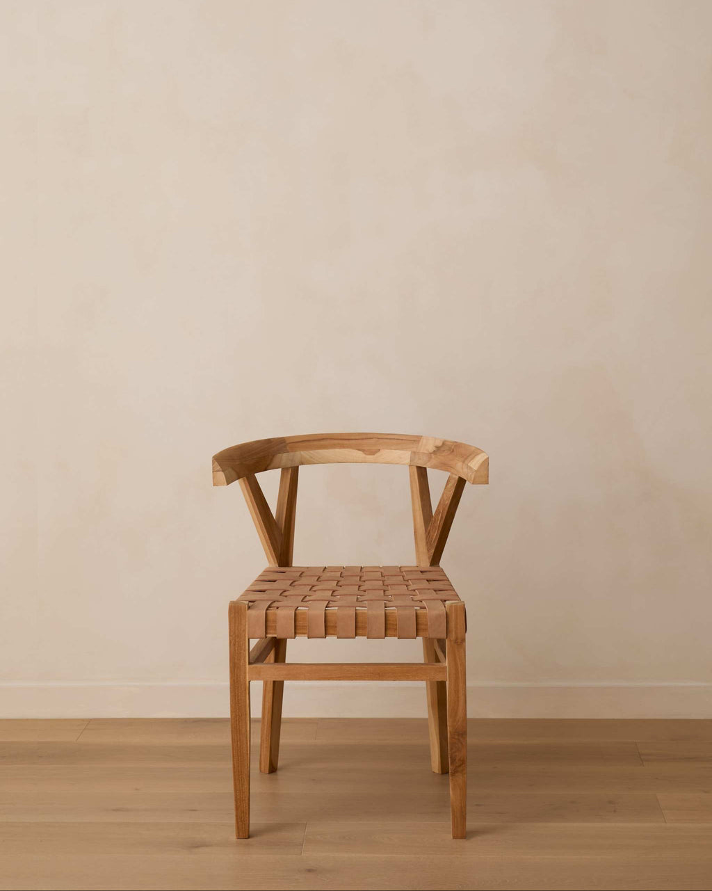 A woven leather wishbone dining chair with a curved backrest and a woven seat, positioned against a neutral beige plaster wall and light wood floor.