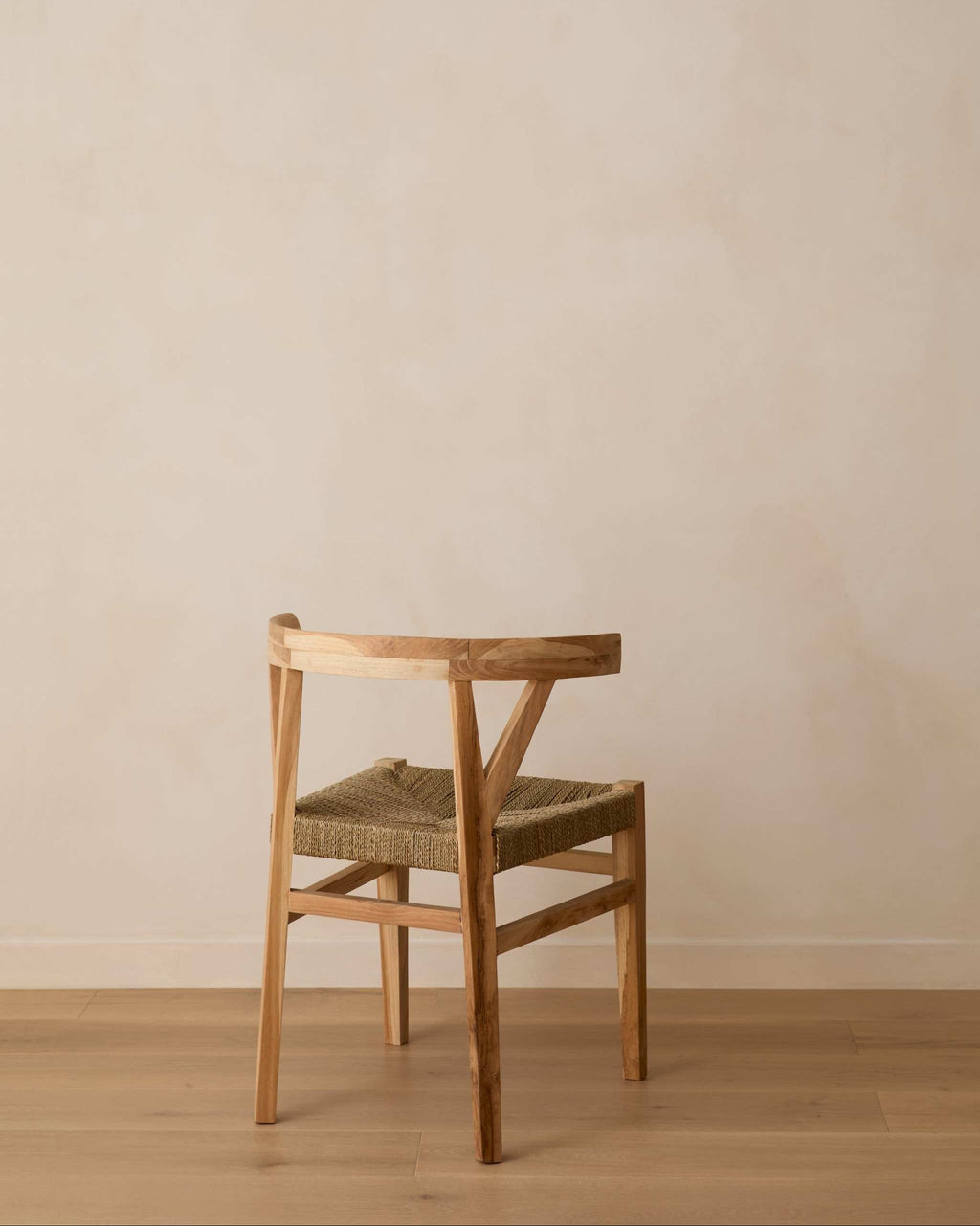 A woven wishbone dining chair with a woven seat, positioned against a minimalist beige plaster wall and light wood floor.