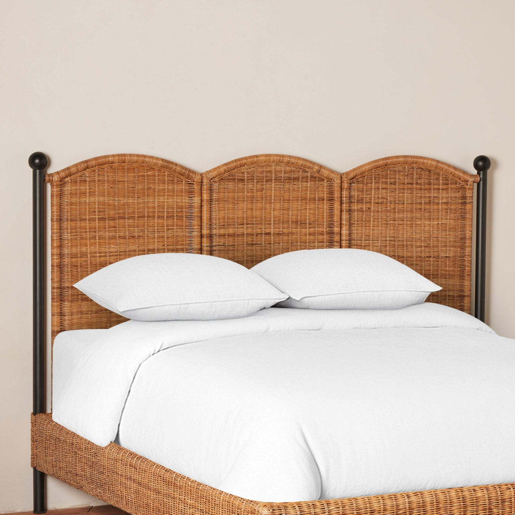 Detail of Lacoste King Bed with scalloped woven rattan headboard, black iron frame, and white bedding against a beige wall.