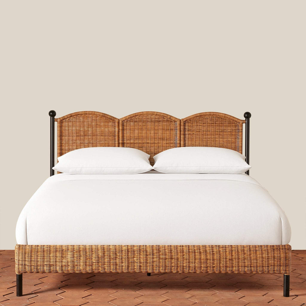 Front view of a woven rattan Lacoste King Bed with a scalloped-shaped headboard on a terracotta tile floor with a beige wall background.