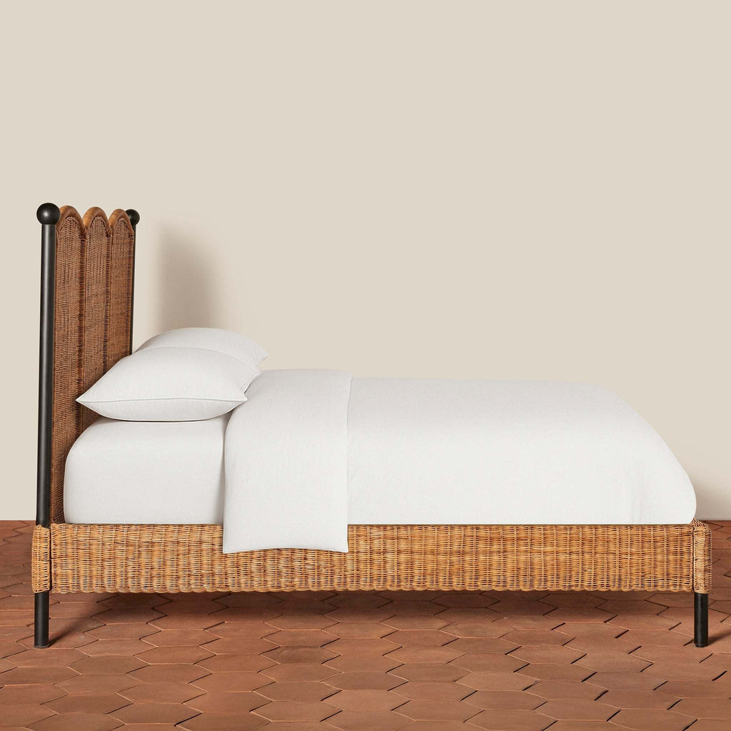Side view of woven rattan Lacoste King Bed with black iron frame and white bedding on a terracotta tile floor against a beige wall.