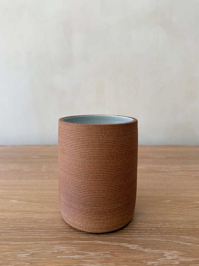 Terracotta ceramic utensil jar with textured exterior and light blue interior on a wooden surface.