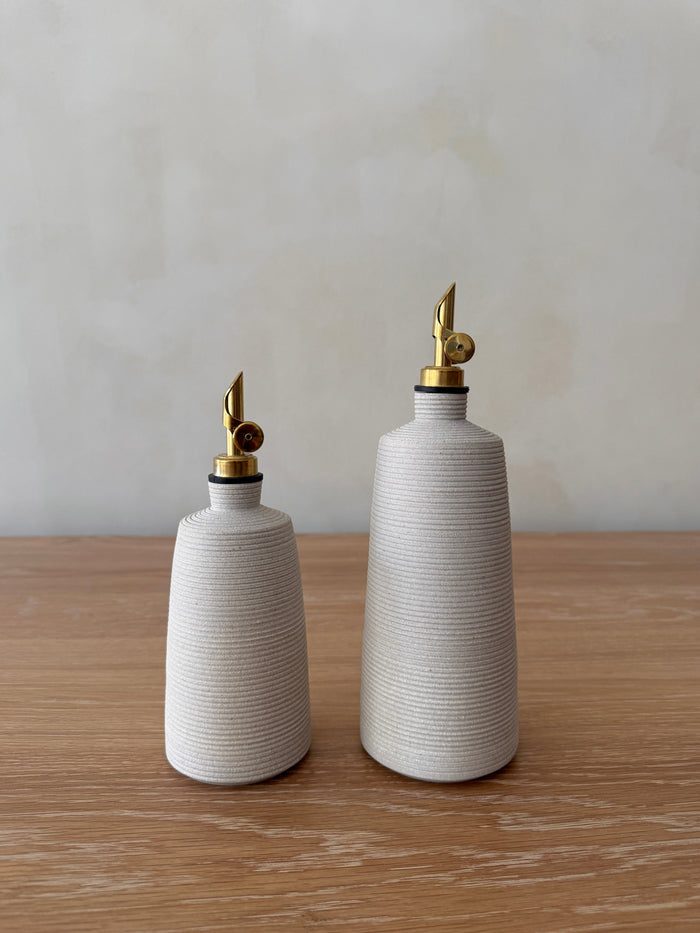 Two white ceramic olive and balsamic bottles with brass nozzles on a wooden surface and light gray background
