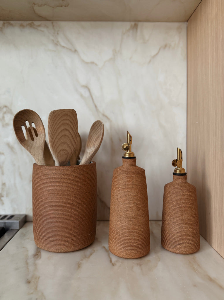 Terracotta Olive oil bottle with balsamic vinegar bottle and utensil jar with wooden spoons on a marble countertop