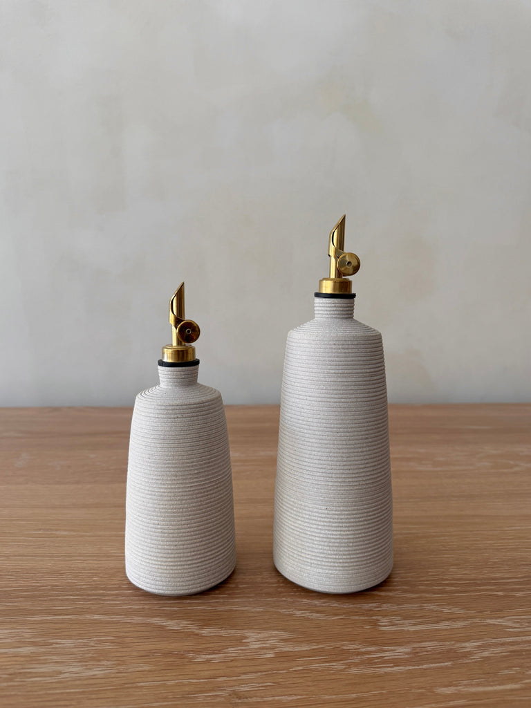 Two white ceramic oil bottles with brass nozzles on a wooden surface and light gray background