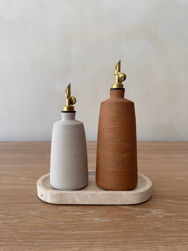 Two ceramic oil dispensers, one white and one terracotta, on a travertine tray with a neutral background.