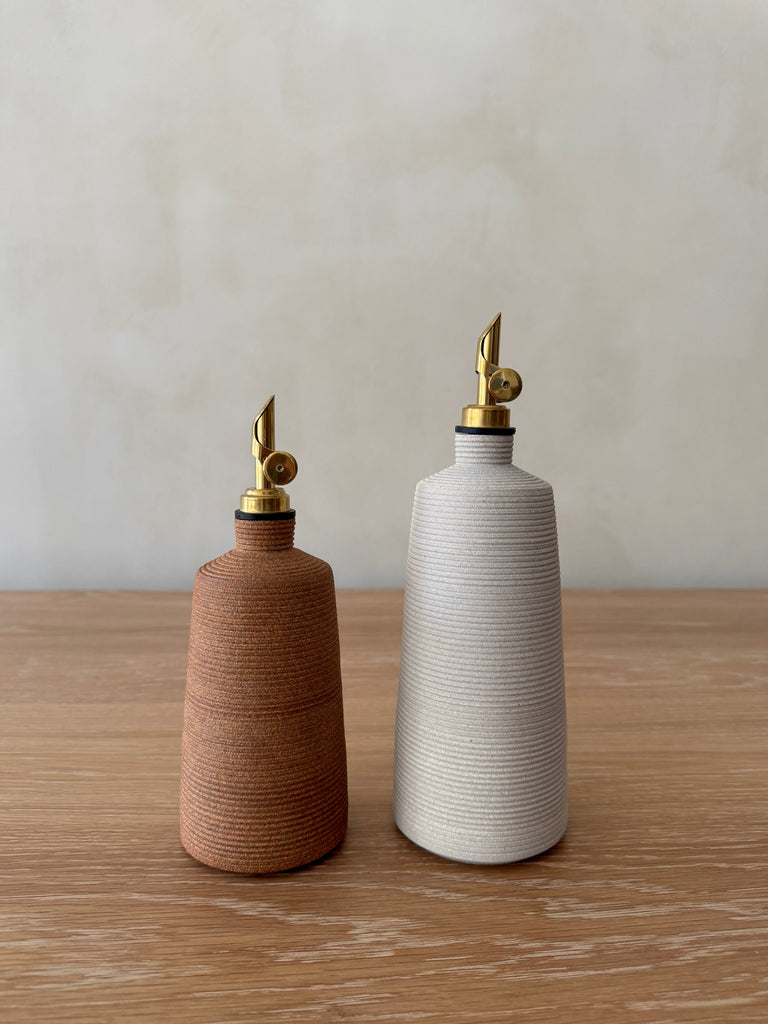 Two ceramic oil dispensers, one brown and one white, on a wooden surface with a neutral background.