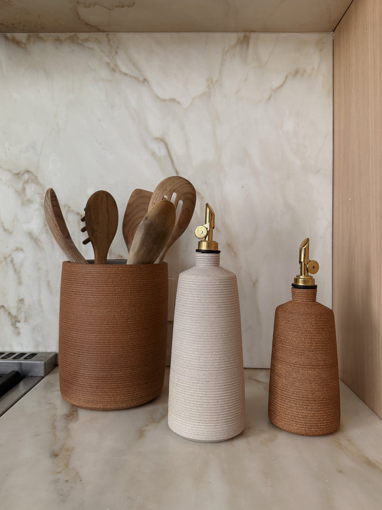 Ceramic utensil jar and oil bottles on a marble countertop with a neutral color palette.