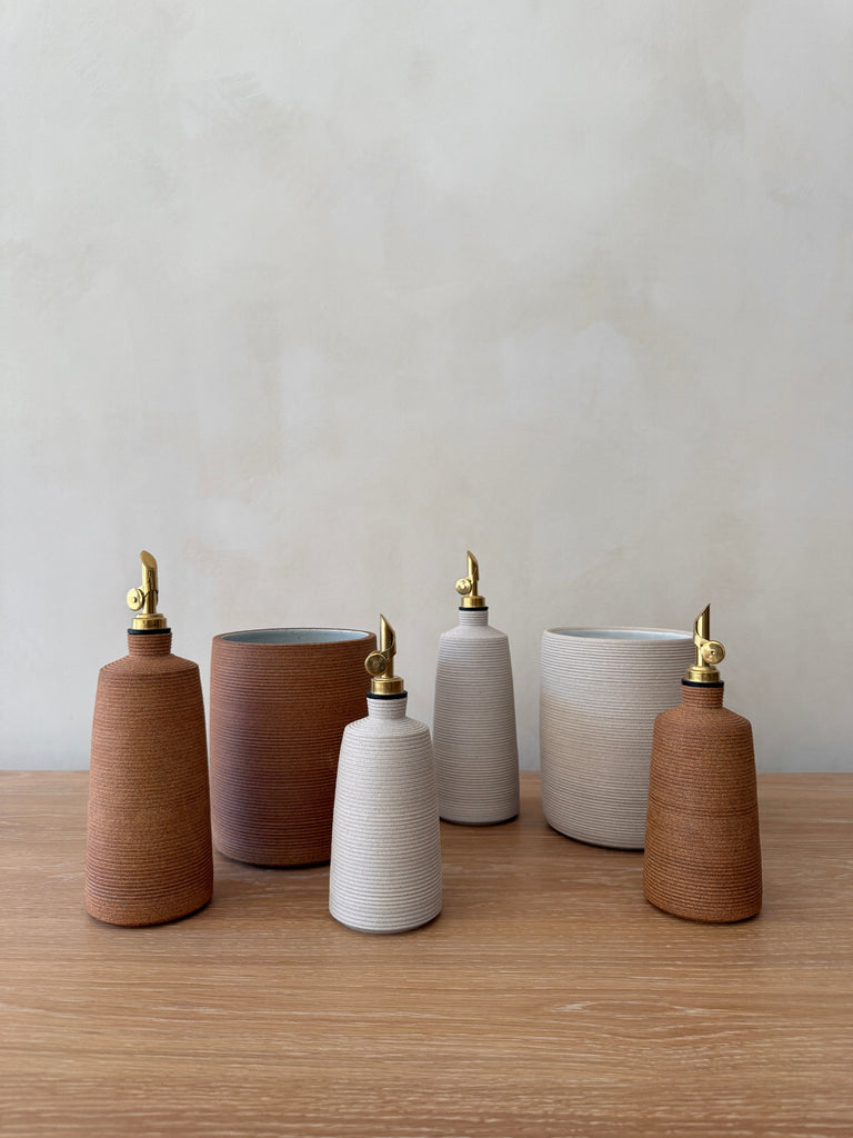 Set of ceramic utensil jars and oil bottles with brass nozzles on a wooden surface.