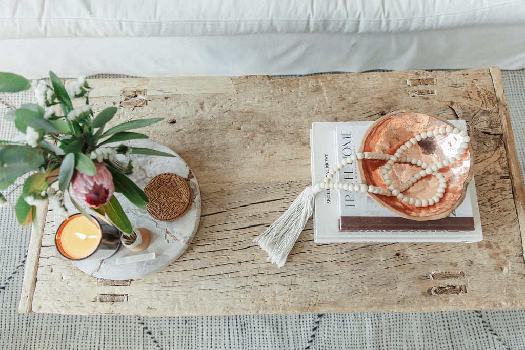 Rustic wooden coffee table with a Travel Home book, decorative beads, a copper bowl, a candle, and a flower arrangement