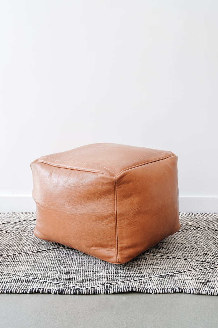 Angled view of Square Leather Moroccan Pouf on a Moroccan Kilim Rug against a white wall. - Saffron and Poe