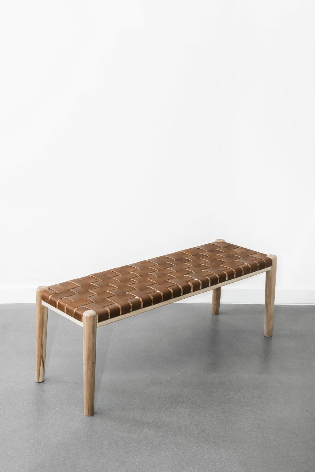 Woven leather bench with teak frame and saddle-colored leather straps on gray floor against white wall