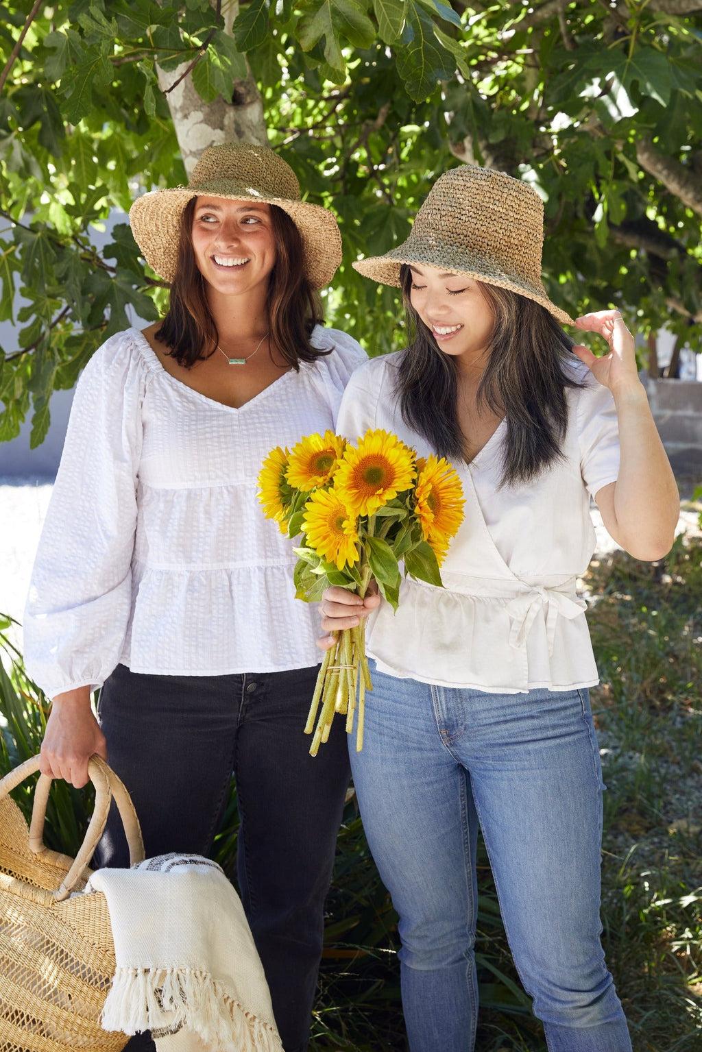 Two women wearing handwoven Balinese bucket hats outdoors, one holding sunflowers and the other carrying a woven bag with a blanket.