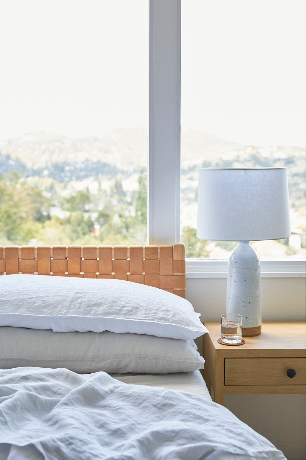 Cozy bed with natural linen sheet set, pillows, wooden headboard, and bedside table with lamp and glass of water near window.
