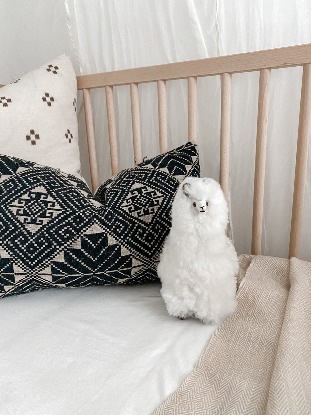 Cute white Alpaquita stuffed animal made of soft baby alpaca wool on a bed with patterned pillows and neutral bedding.