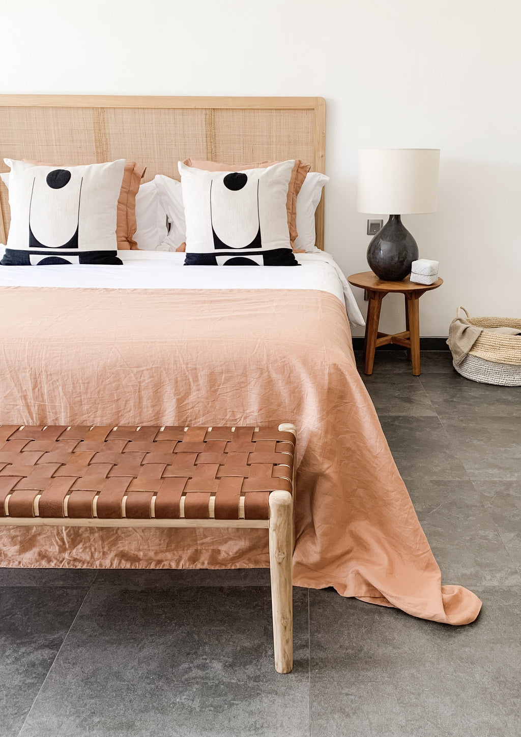 Collapsible woven hyacinth basket placed beside a bedside table in a modern bedroom with peach bedding and decorative pillows