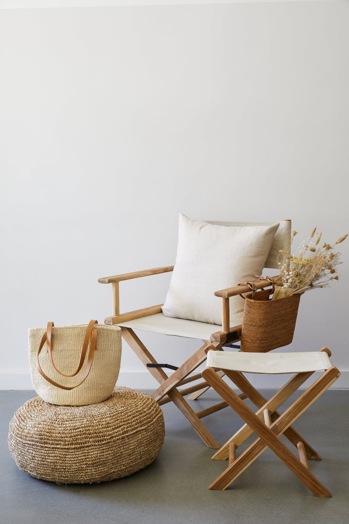 Canvas Director's Chair in Oatmeal with Ivory Director's Pillow, Oatmeal Canvas Folding Stool, Tenganan Side Basket, Natural Woven Pouf, and Woven Sisal and Leather Tote against a white background and concrete flooring. - Saffron and Poe