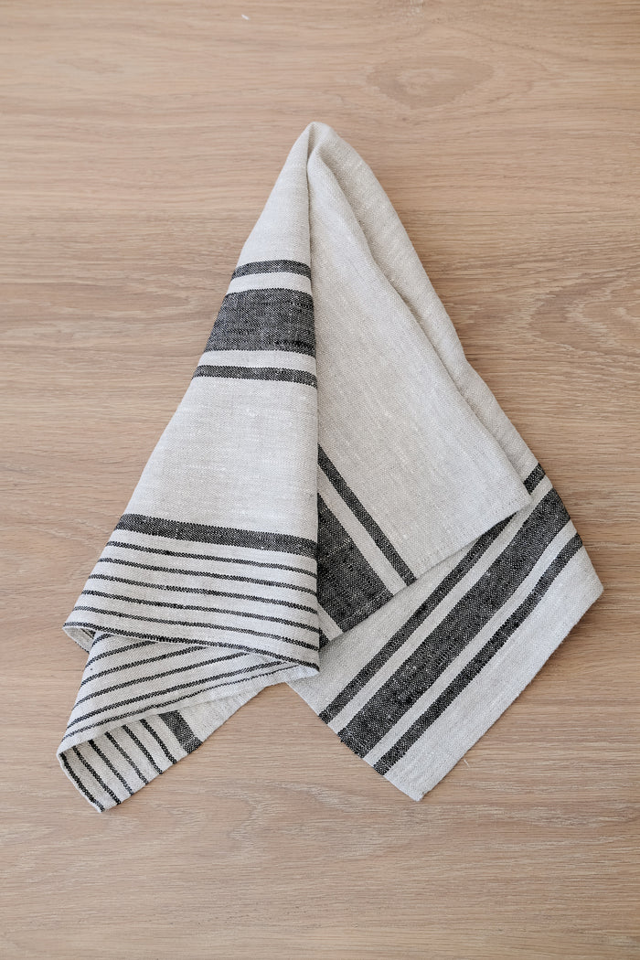 Top down view of draped Dark Grey Stripe Linen Hand Towel (Set of 2) on a White Oak Dining Table. - Saffron and Poe