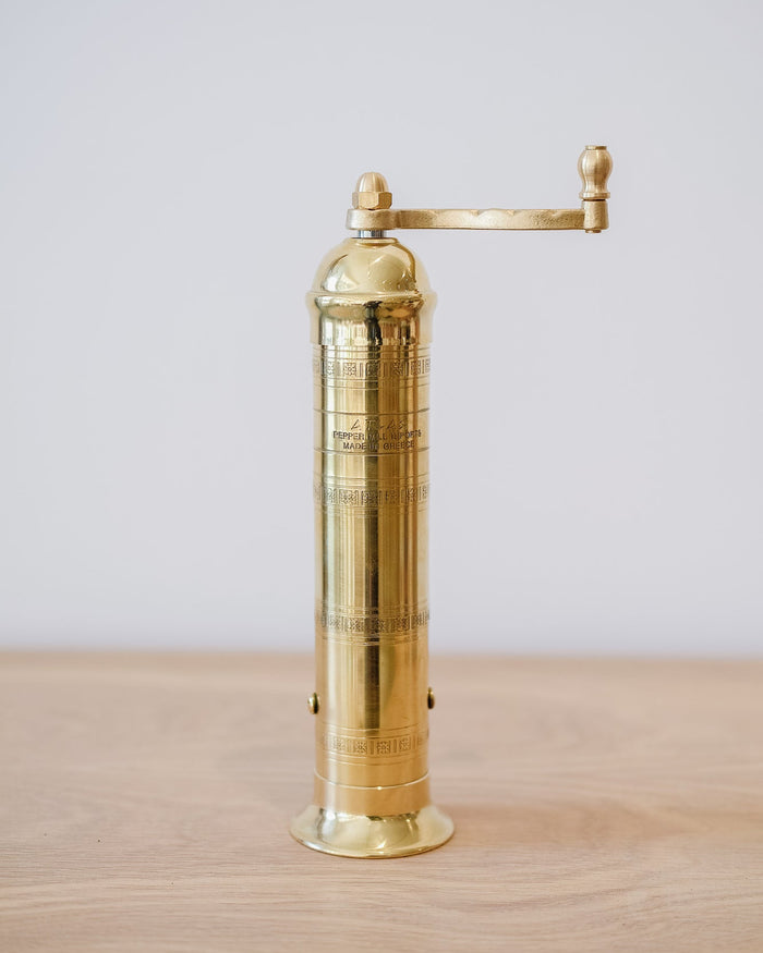 Front view of Atlas Brass Pepper Grinder - 9" on an oak table against a white wall.