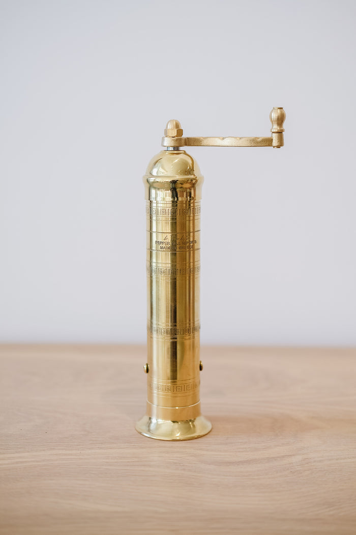 Front view of Atlas Brass Pepper Grinder - 9" on an oak table against a white wall. - Saffron and Poe