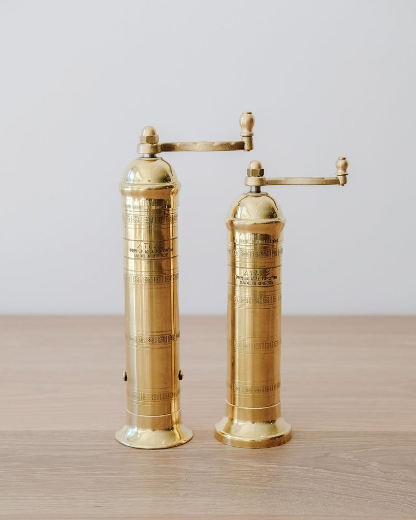 Atlas Brass Salt and Pepper Grinders on an oak table against a white wall.