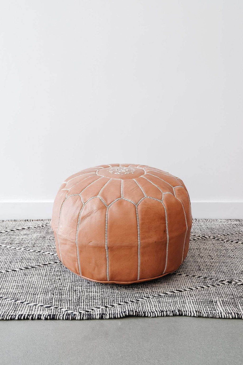 Embroidered Moroccan leather pouf ottoman on gray patterned rug against white wall