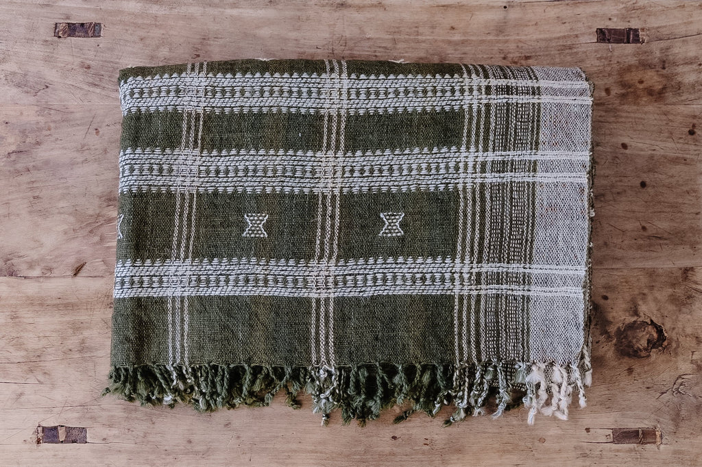 Handwoven Bhujodi Blanket in olive green with white pastoral motifs and fringed edges on wooden surface