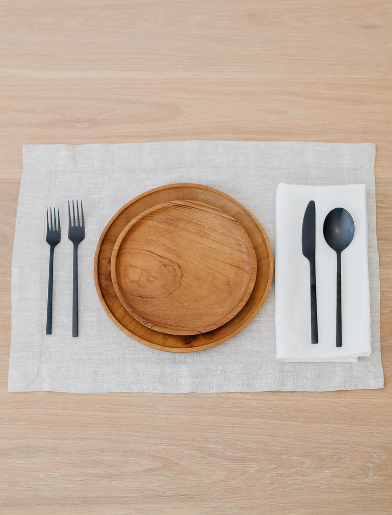 Front view of small teak plate and large teak plate on linen placemat with black utensils and ivory linen napkin. - Saffron and Poe. 