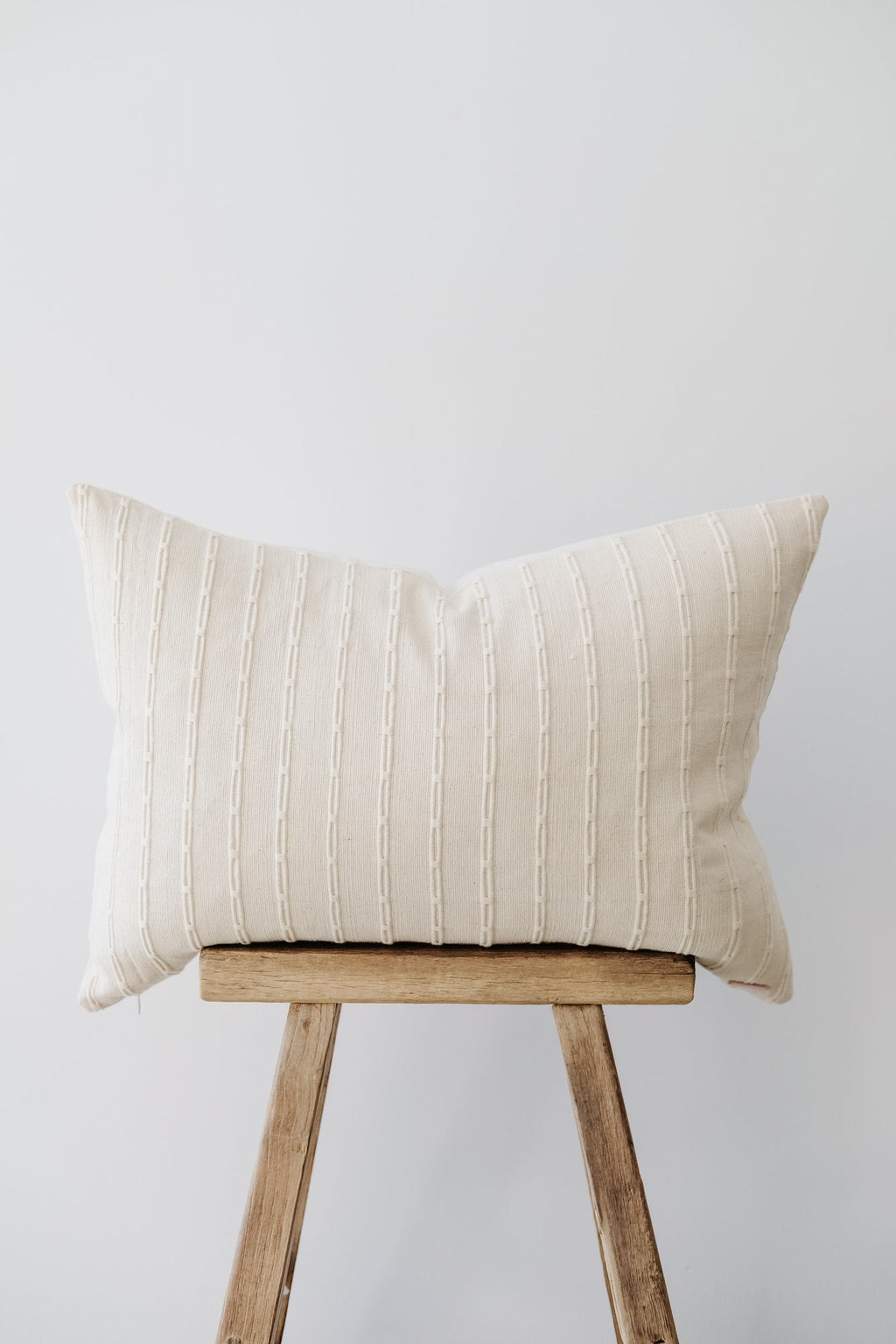 Ivory embroidered pillow with subtle textured pattern on wooden stool against white background