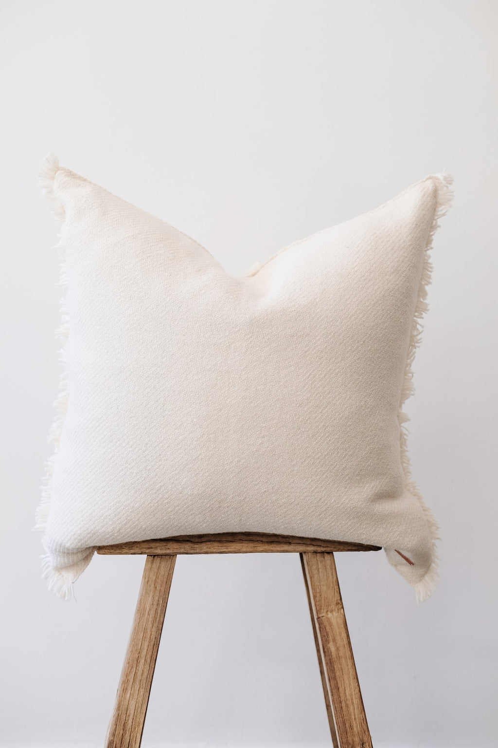 Ivory baby alpaca pillow with fringe on wooden stool against white background