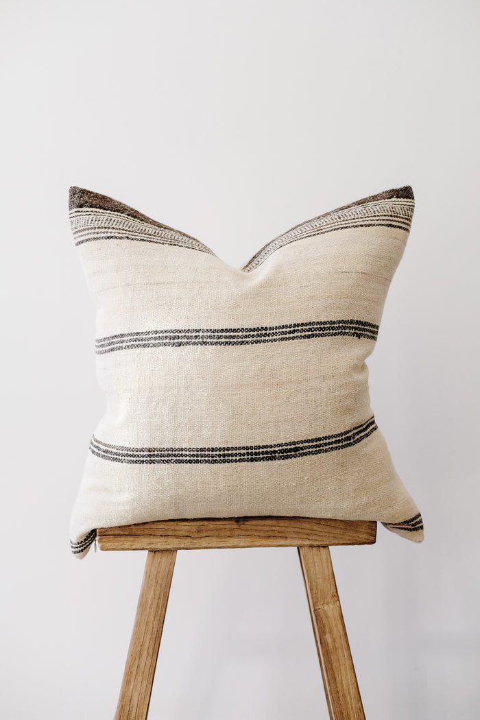 Front view of No. 31 - Handwoven Bhujodi Pillow - Natural on an Antique Stool against a white wall. - Saffron and Poe