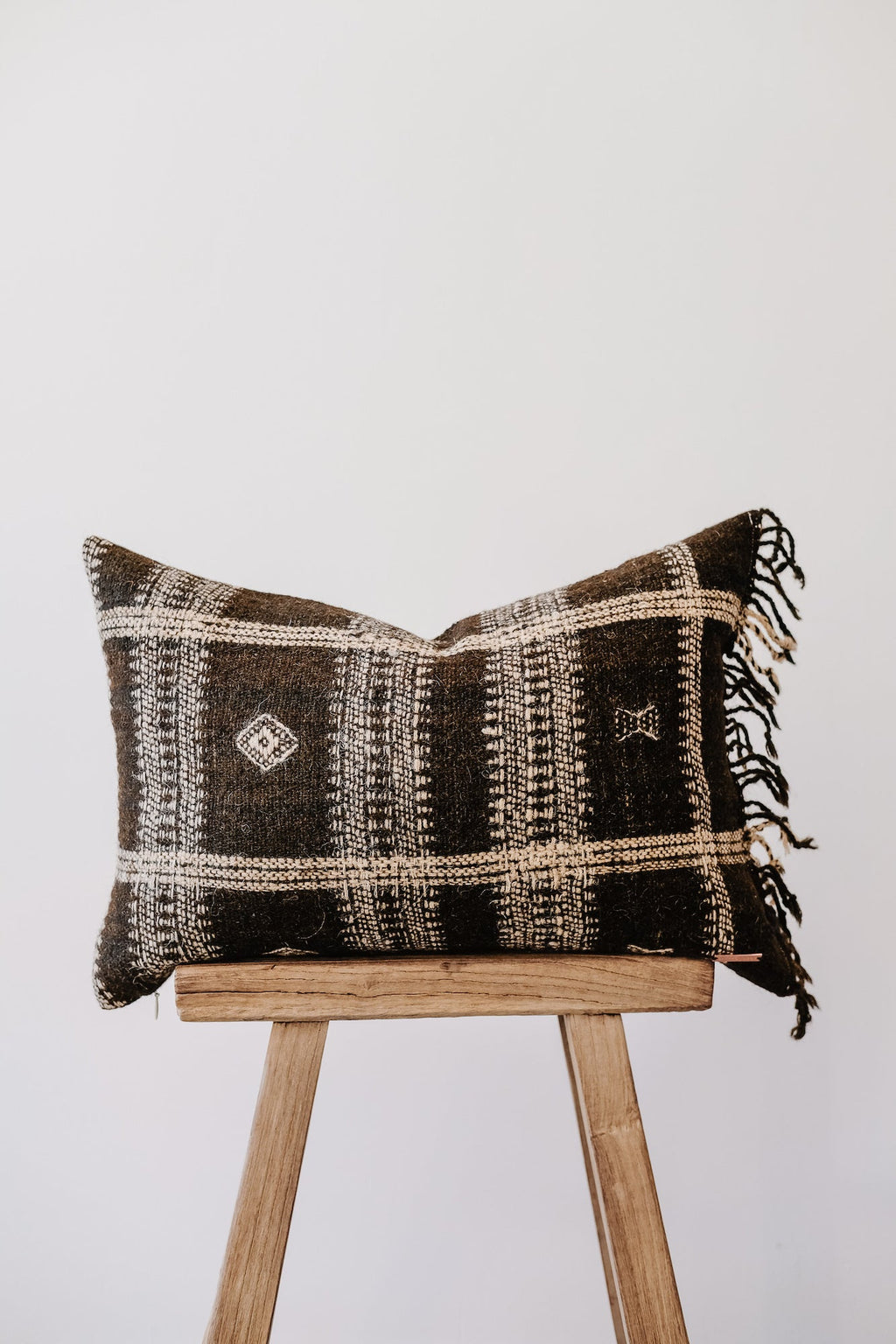 Handwoven Bhujodi pillow with black and white geometric patterns and fringe on wooden stool against white background