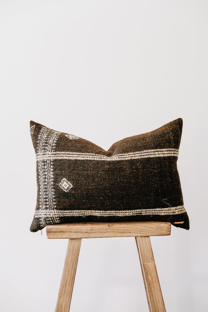 Front view of No. 39 - Handwoven Bhujodi Lumbar Pillow - Dark Brown on an Antique Stool against a white wall. - Saffron and Poe