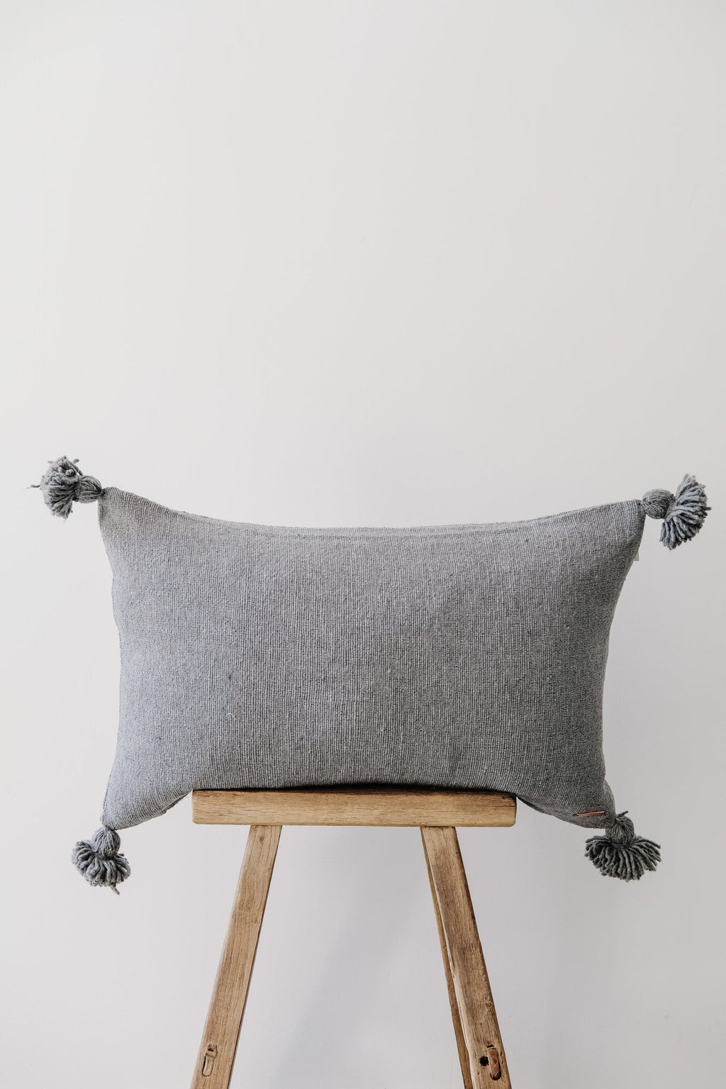 Grey Moroccan pom pillow with tassels on wooden stool against white background